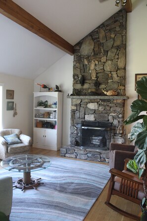 A living room with a stone fireplace, wooden beams, a white shelving unit, and a glass coffee table. - Serene 4-bedroom house with water access in charming Phippsburg (Phippsburg)