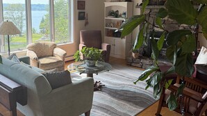A living room with a stone fireplace, large windows, a sofa, a chair, and a table with a plant. - Serene 4-bedroom house with water access in charming Phippsburg (Phippsburg)