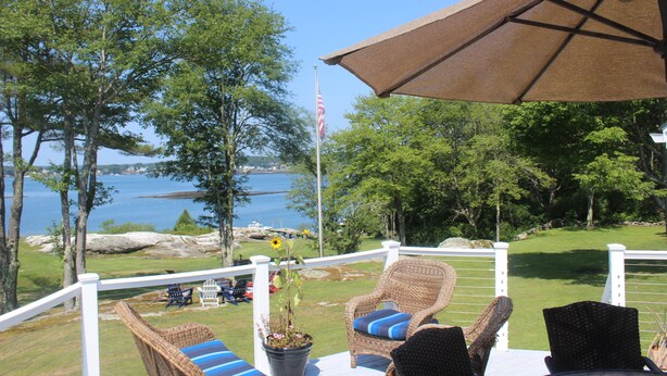 A deck with wicker chairs and a table, overlooking a body of water and trees. - Serene 4-bedroom house with water access in charming Phippsburg (Phippsburg)