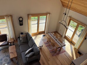 Un salon chaleureux avec un canapĂ©, une petite table et une table Ă manger avec des chaises. De grandes fenĂȘtres sont ornĂ©es de rideaux, une horloge est accrochĂ©e au mur et le plafond est dĂ©corĂ© de poutres en bois.