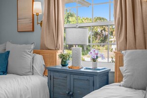 A bedroom with two beds, a bedside table with a lamp, a framed picture, and a window with curtains. - Waterfront Oasis - Heated Pool, Kayaks + Game Room (Palm Coast)
