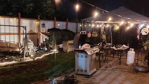 Un patio trasero con una parrilla, luces colgantes y un espacio cubierto para sentarse.