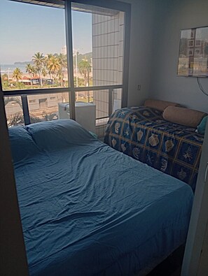 A bedroom with a large window, a bed with a patterned blanket, and a view of palm trees and buildings. - Beachfront property with two oceanfront balconies (São Vicente)