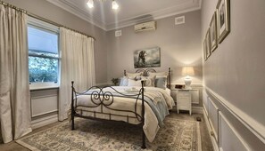 A bedroom with a four-poster bed, a nightstand with a lamp, a painting on the wall, and a window with curtains.