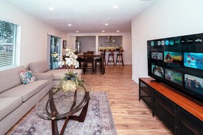 Uma sala de estar moderna com um sofá, uma mesa de centro com uma planta, uma área de jantar com mesa e cadeiras, e uma TV exibindo serviços de streaming.