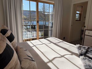 A bedroom with a large window, a bed, a dresser, and a view of a lake and trees.