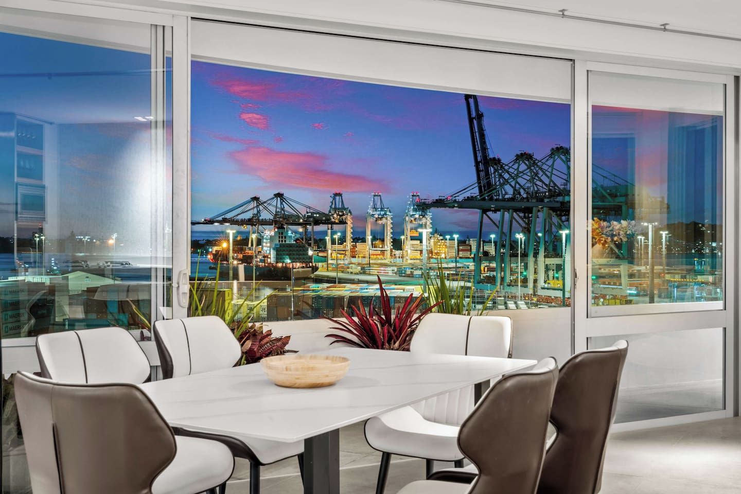 Un comedor moderno con una mesa grande y sillas, ventanas amplias con vistas a un puerto y grúas, y una barandilla de vidrio.