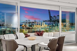 Ein moderner Essbereich mit einem großen Tisch, weißen Stühlen und Blick auf einen Hafen mit Kränen und Schiffen.