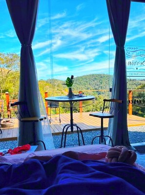 A bed with a view of a deck, a table with chairs, and a vase with a plant. - Aysú Mirante Container (Paty do Alferes)