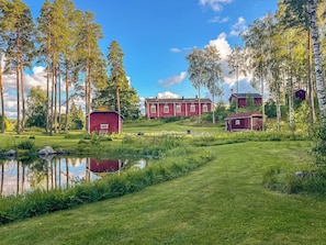 Eine friedliche Landschaft mit Teich, roten Scheunen und einem großen Haus mit vielen Fenstern.