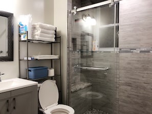 Un baño moderno con ducha envuelta en vidrio, inodoro blanco y repisa con toallas y artículos de aseo.