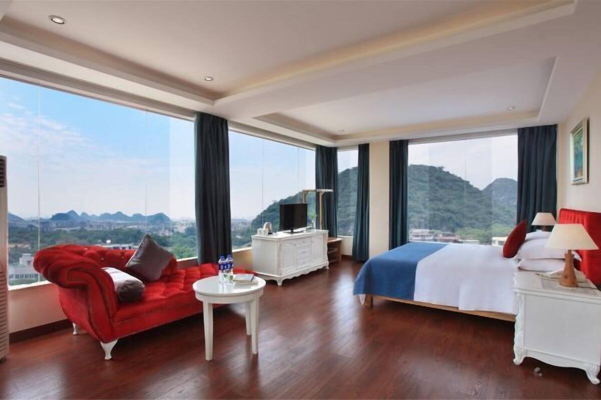 A hotel room with a bed, a red sofa, a white side table, a TV, and a large window with a mountain view.