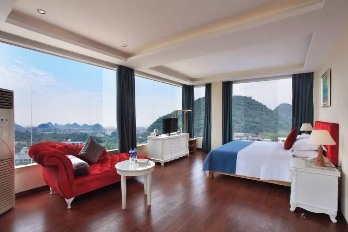 A hotel room with a bed, a red sofa, a white dresser, and a large window offering a mountain view.