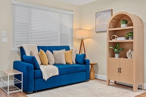 Ein blaues Sofa mit Kissen, ein kleiner weißer Beistelltisch, eine Stehlampe und eine Holzkommode mit Regalen.