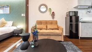 A modern living room with a sofa, a coffee table, a small side table, a decorative mirror, and a kitchen area with a refrigerator and microwave. - FDS Panache (OSAKA/NISHINARIKU/HANAZONOMINAMI)