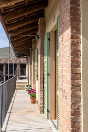 A covered balcony with brick walls and green shutters, potted plants, and a railing.
