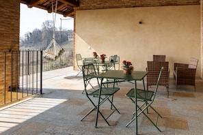 A covered outdoor patio with a table and chairs, a hanging macrame light, and a view of the countryside.