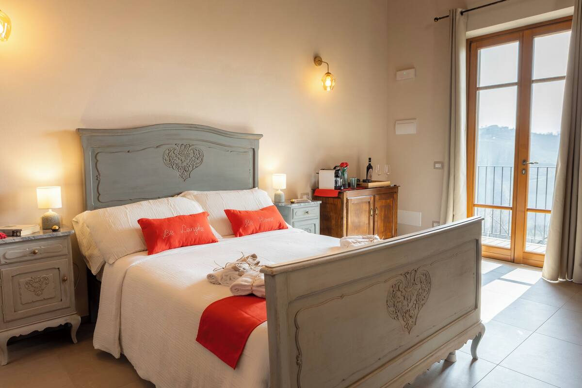 Una cama bien hecha con ropa de cama blanca y roja, una mesita de noche con una lámpara y vistas al exterior a través de una puerta de cristal.