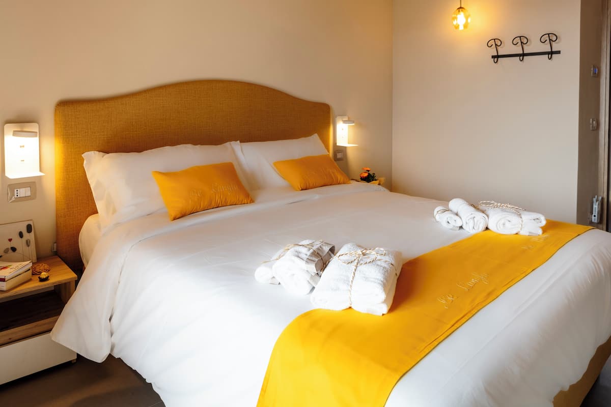 Una cama bien tendida con ropa de cama blanca y amarilla, una mesita de noche con una lámpara y un aplique en la pared.