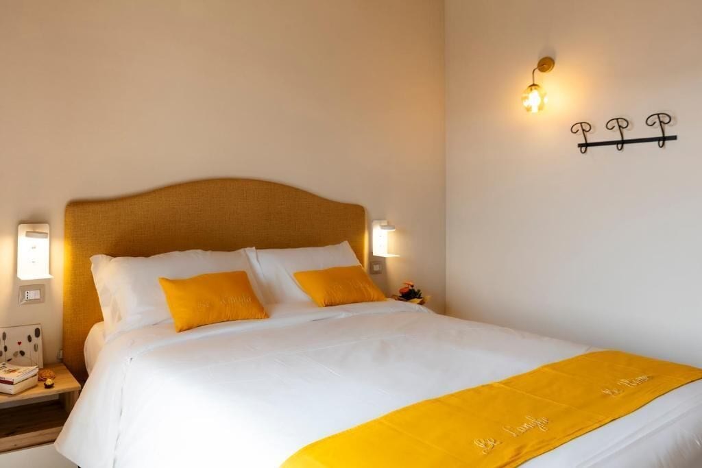 A neatly made bed with white and yellow bedding, a bedside table with a book and a small decorative item, and a wall-mounted light fixture.