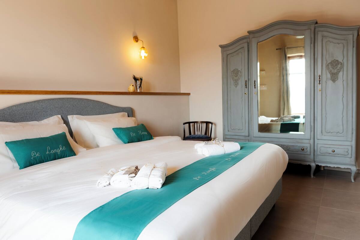 Una cama bien hecha con un corredor azul turquesa y almohadas, una mesita de noche de madera, un espejo grande y una lámpara de pared.