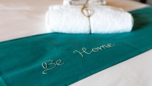 A neatly folded white towel with a gold string tied around it, placed on a bed with a teal bedspread that has 'Be Home' embroidered on it. - Be Langhe in Monforte (Monforte d'Alba)