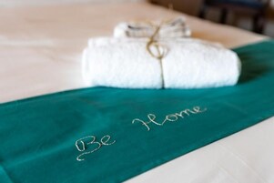 Una toalla blanca doblada con un cordón dorado atado, colocada sobre una cama con un edredón color turquesa que tiene bordado el texto 'Be Home'.