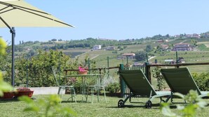 A patio with a table and chairs, a wheelbarrow, and a view of rolling hills and buildings. - Be Langhe in Monforte (Monforte d'Alba)
