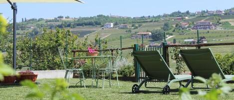 Une terrasse avec une table et des chaises, une brouette, et une vue sur des collines ondoyantes et des bâtiments.