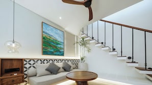 A modern living room with a sofa, a coffee table, a painting, and a staircase. - Nuho Living (Pecatu)