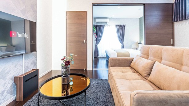 A modern hotel room with a sofa, a coffee table with a vase, a TV, and a door leading to a balcony. - FDS Luxe (OSAKA/NISHINARIKU/HANAZONOMINAMI)
