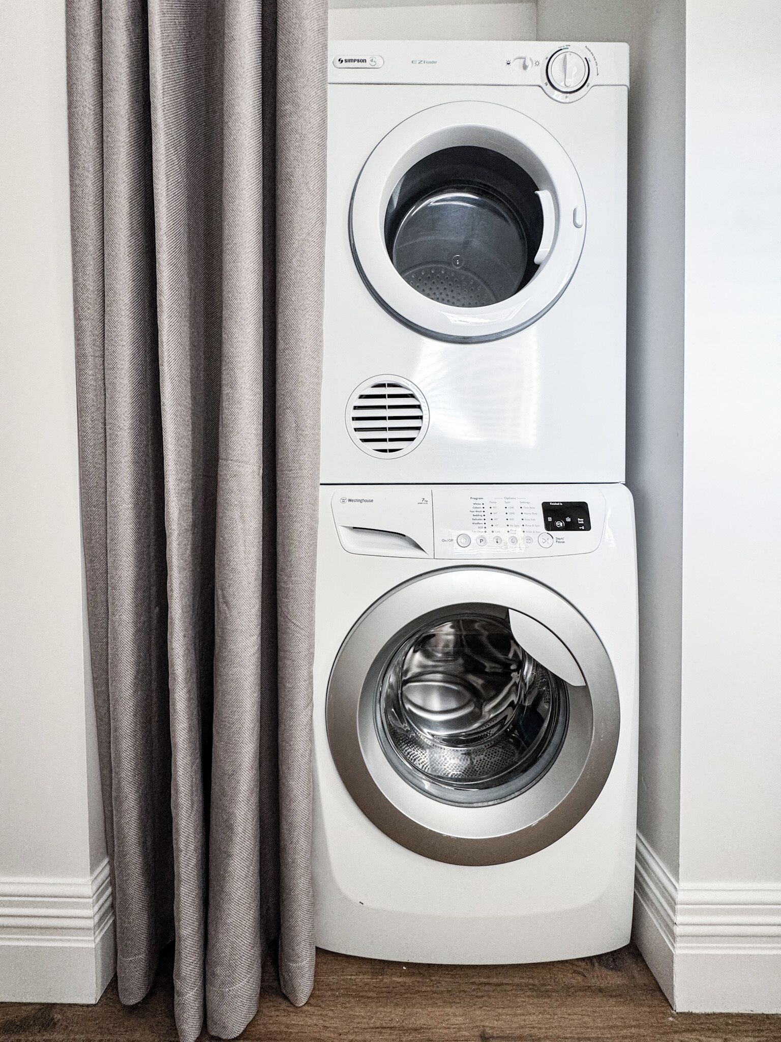 Een wasmachine en droger, gestapeld in een hoek, met een houten vloer en een gordijn.