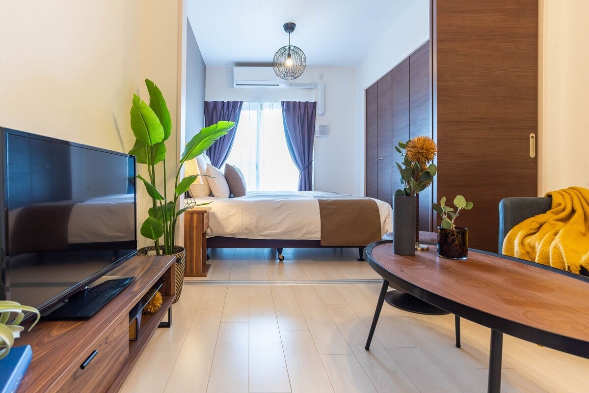 A modern hotel room with a bed, a TV, a dining table, and a plant.