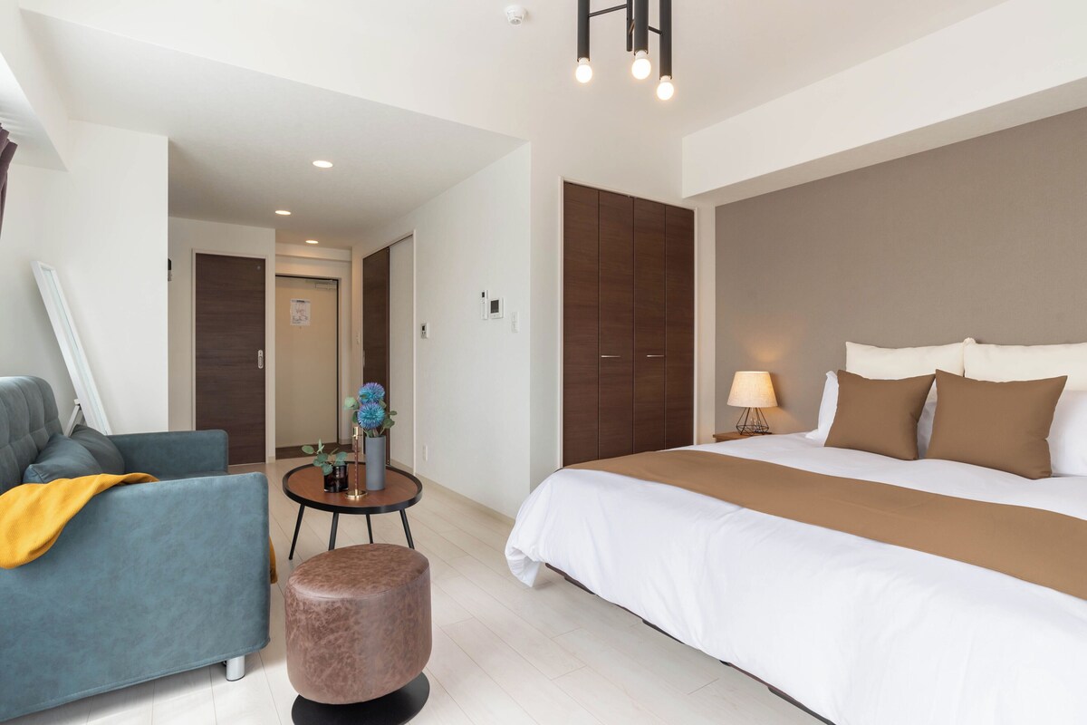 A modern hotel room with a bed, a sofa, a small table with a lamp, and a round ottoman.