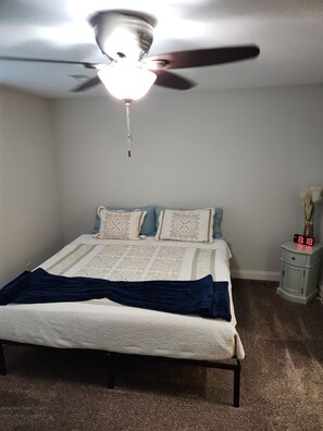Ein Schlafzimmer mit einem Bett, einem Deckenventilator, einem Nachttisch mit einer Uhr und einem Teppichboden.