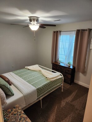 Un dormitorio con cama, ventilador de techo, ventana con cortinas y cómoda.