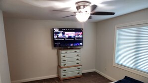 Une chambre dotée d’un ventilateur de plafond, d’un téléviseur fixé au mur, d’une commode avec des tiroirs, d’un lit recouvert d’une couverture bleue et d’une fenêtre avec des stores.