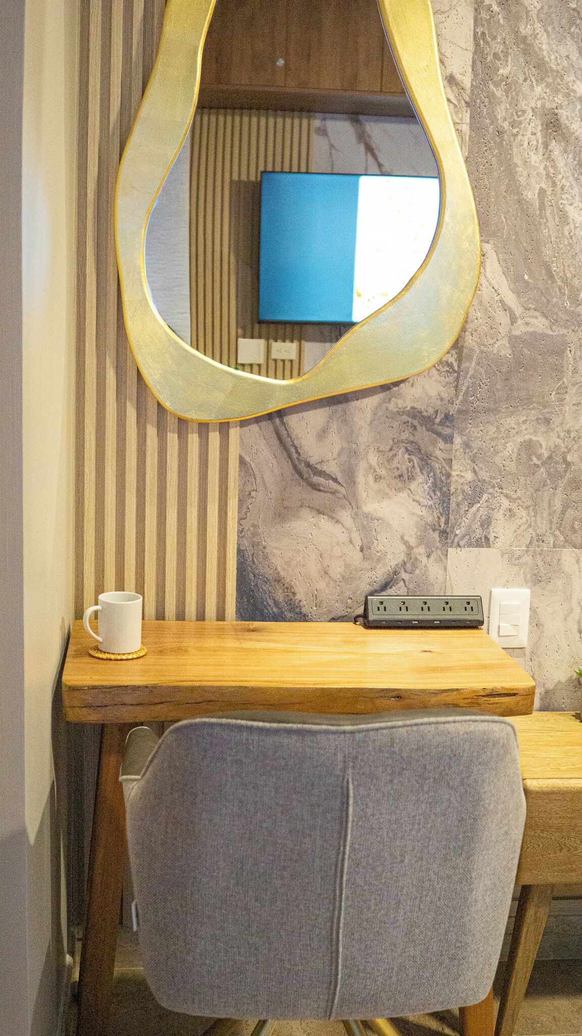 A wooden desk with a mirror, a white mug, and a power outlet.