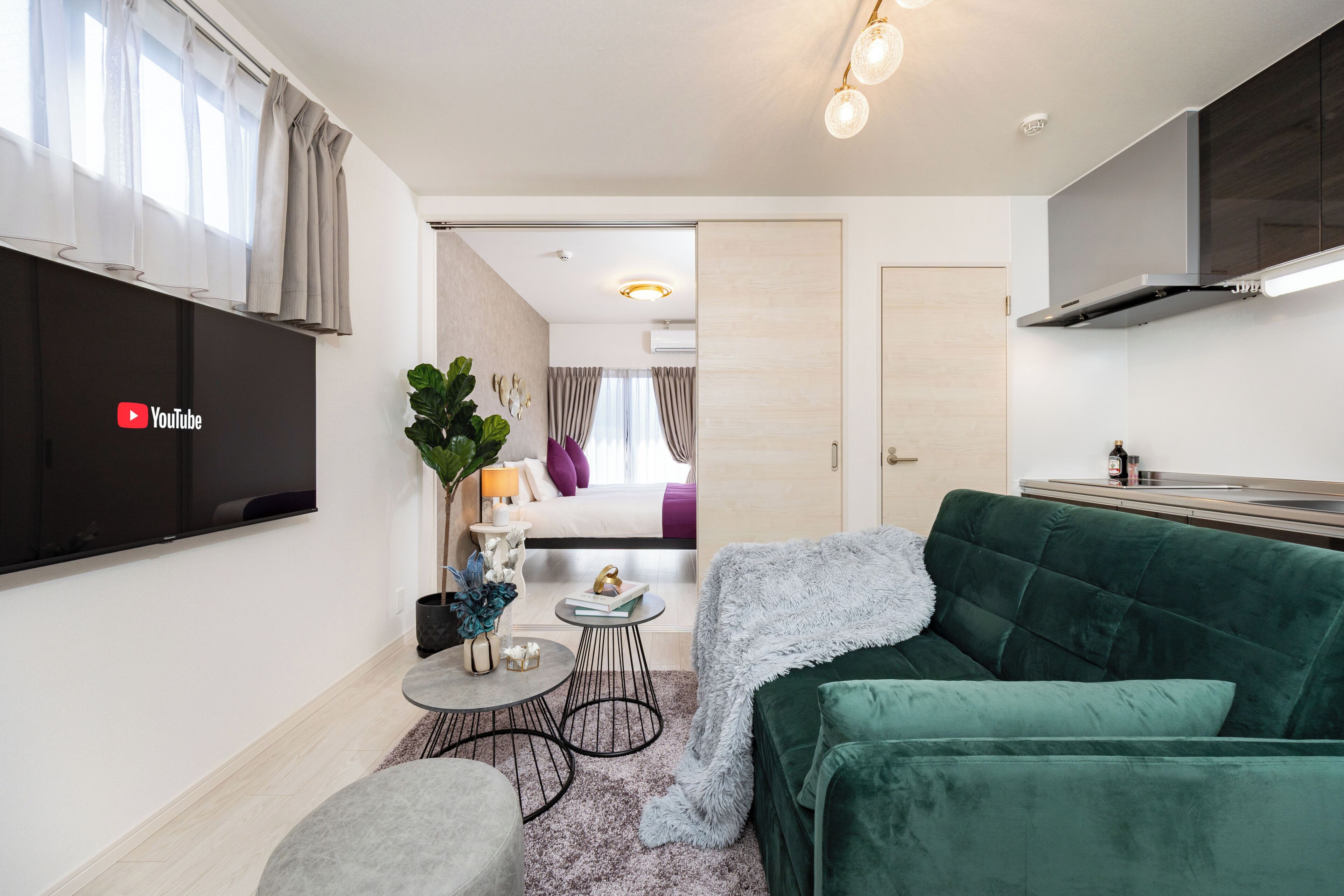 A compact living space with a flat-screen TV, a green sofa, a small round table, and a bed with a purple accent.