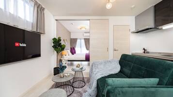 A compact living space with a flat-screen TV, a green sofa, a small round table, and a bed with a purple accent.