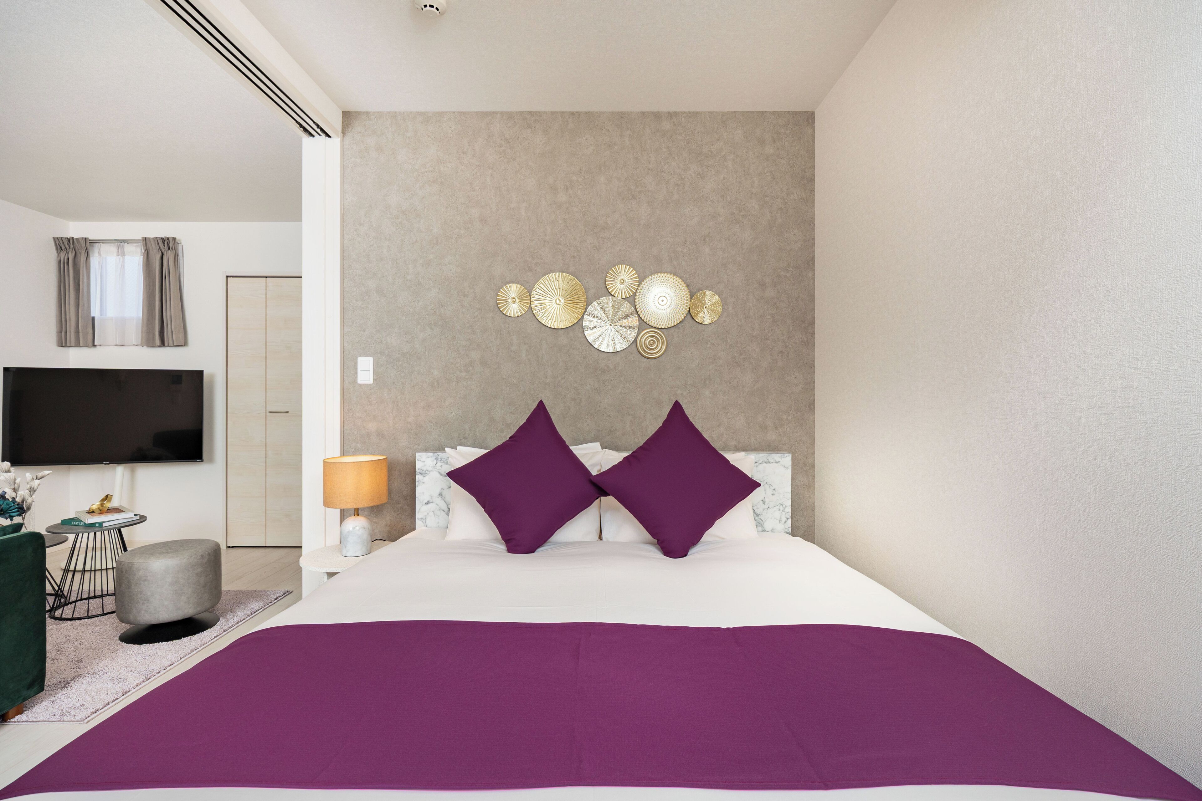A modern bedroom with a large bed, a flat-screen TV, a bedside table with a lamp, and a decorative wall piece.