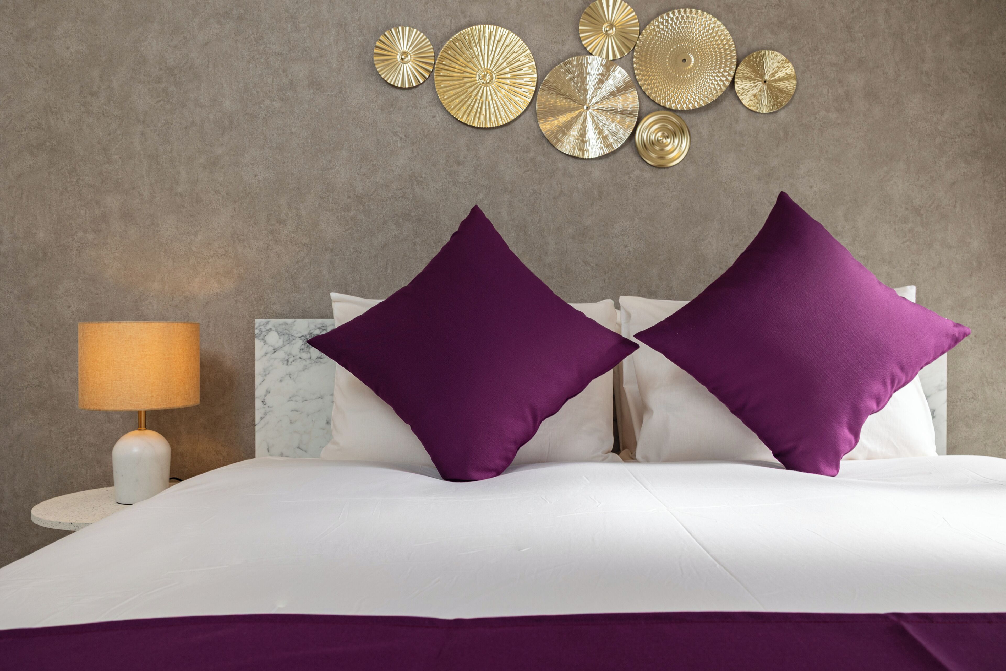 A bed with white bedding and two purple pillows, a round table with a lamp, and decorative wall art.
