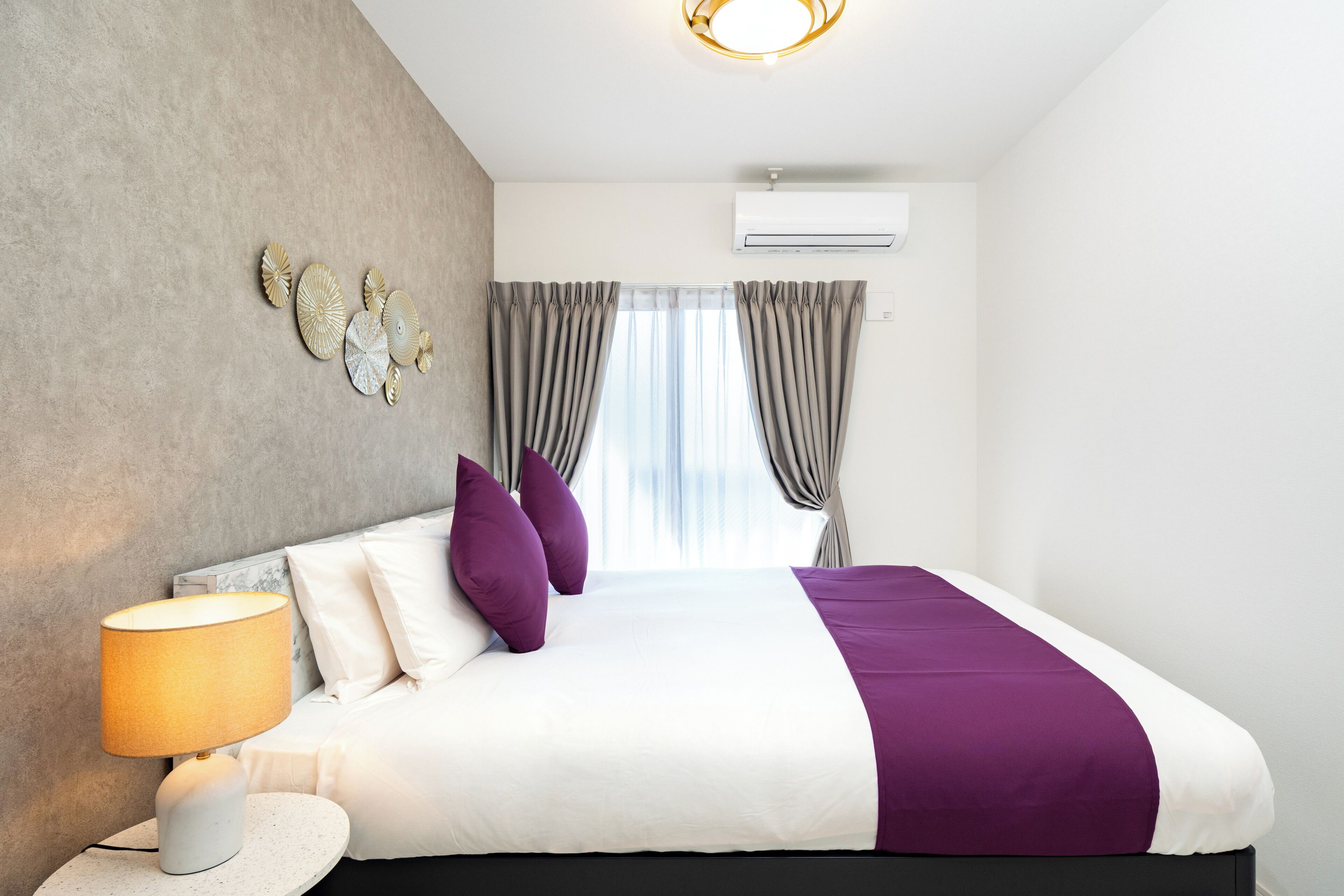 A modern hotel room with a bed, a round nightstand, a lamp, a wall-mounted air conditioner, and a window with curtains.