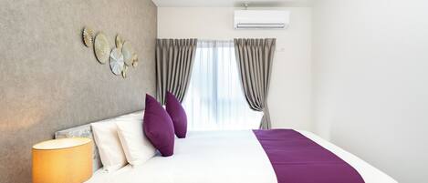 A modern hotel room with a bed, a round nightstand, a lamp, a wall-mounted air conditioner, and a window with curtains.
