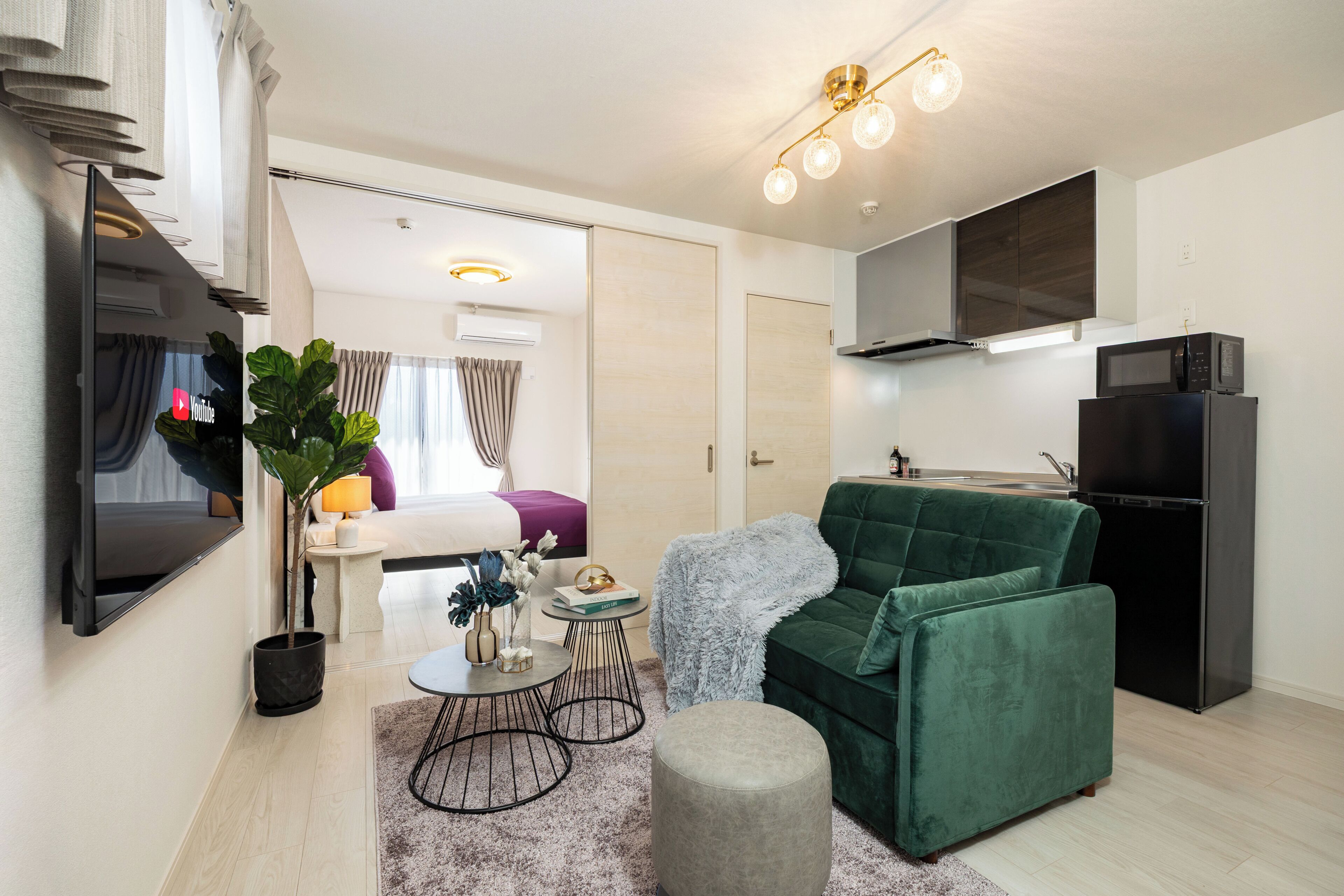 A modern living room with a flat-screen TV, a green sofa, a small round table, and a kitchen area with a microwave and refrigerator.