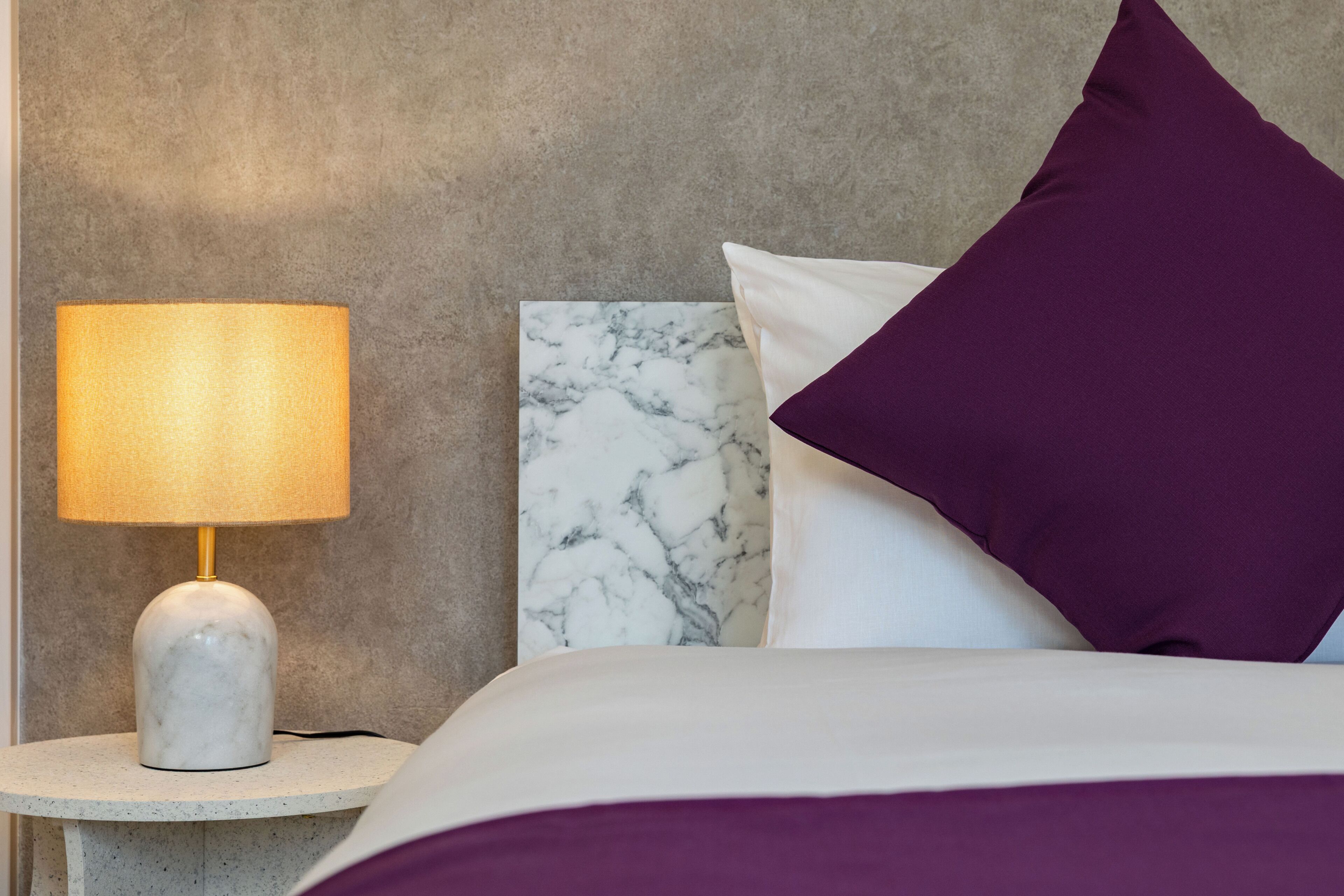 Ein Bett mit einem Marmorkopfstück, ein runder Nachttisch mit einer Lampe und ein violettes Kissen.