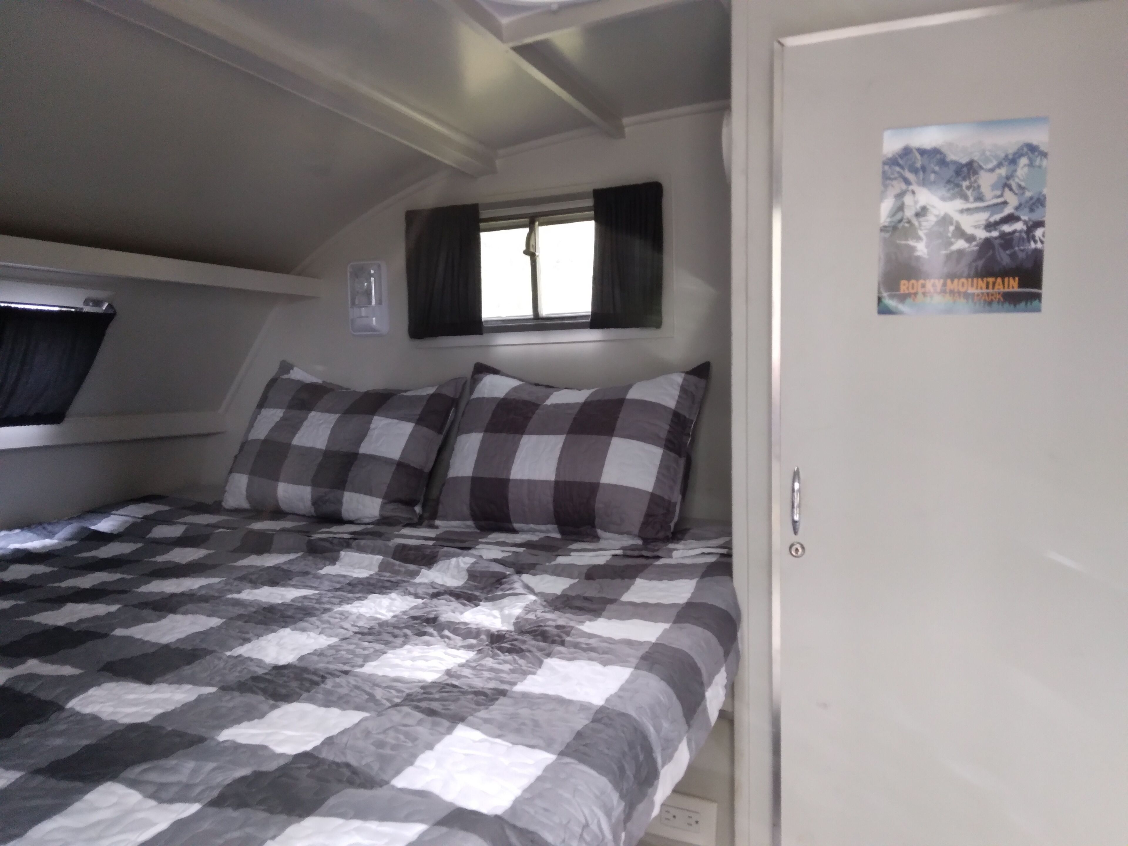 A bedroom with a checkered bedspread, a window with curtains, a poster on the wall, and a door.. Denali Scotty @ Creekside Campground