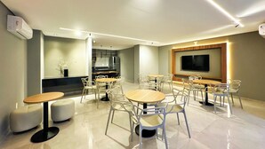 A modern dining area with multiple tables and chairs, a flat-screen TV mounted on the wall, and a kitchen area in the background. - Edifício HIGH (Maceió)