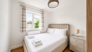 Ein Schlafzimmer mit einem Bett, einem Nachttisch mit Lampe, einem Fenster mit Vorhängen und Blick auf Grün außerhalb.