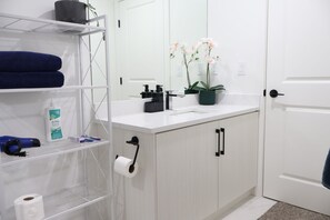 A bathroom with a white sink, a mirror, a towel rack with blue towels, a shelf with toiletries, and a potted orchid plant. - Modern Suite in Seton - King Bed, 85" TV, Near South Health Campus & YMCA (Calgary)